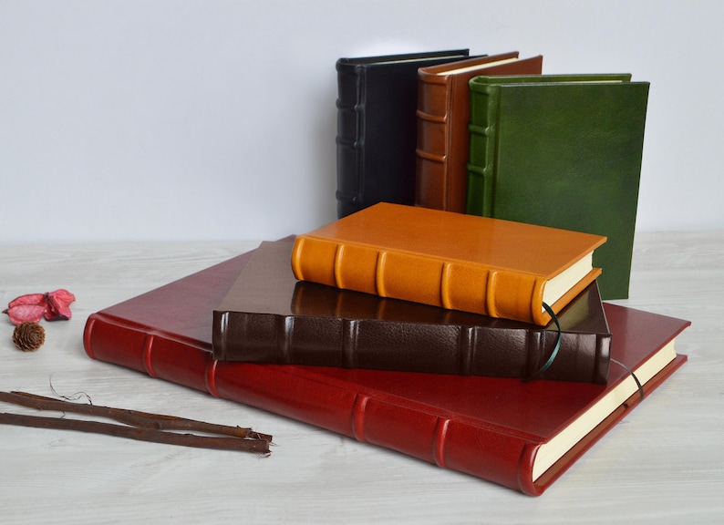 May include: A stack of five leather-bound journals in various colors, including red, brown, orange, green, and black. The journals have decorative raised lines and are arranged in a pyramid shape.