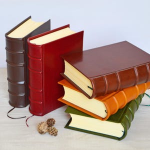 May include: Five hardcover books with smooth leather-like covers in dark brown, red, tan, orange, and olive green. Two books stand upright while three are stacked horizontally. Each book features raised bands on the spine and cream-colored pages.