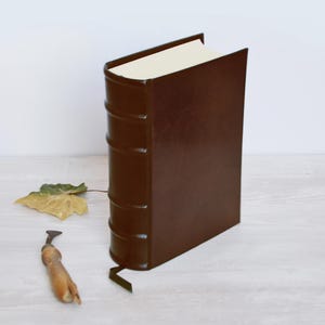 May include: A dark brown, leather-bound book standing upright with a rounded spine featuring five raised bands. The book has cream-colored pages and a thin ribbon bookmark extending from the bottom. A small, dried leaf and a metal tool are nearby.