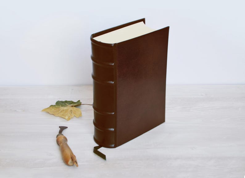 May include: A dark brown, leather-bound book standing upright with a rounded spine featuring five raised bands. The book has cream-colored pages and a thin ribbon bookmark extending from the bottom. A small, dried leaf and a metal tool are nearby.