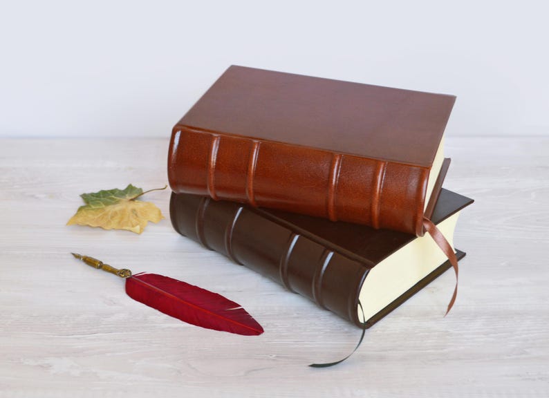 May include: Two hardcover books with smooth, solid-colored covers in shades of medium brown and dark brown, featuring raised bands on the spines and ribbon bookmarks. A single red feather quill pen with a metallic tip lies beside the books.
