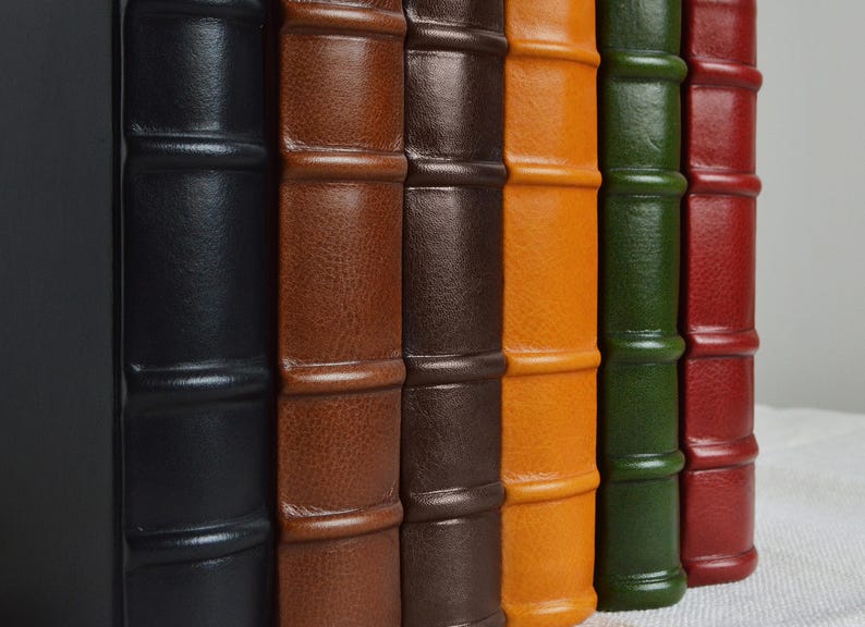 May include: A row of six vertical book spines featuring raised bands and a textured finish. The colors from left to right are black, light brown, dark brown, yellow-orange, forest green, and deep red, all standing upright in a side-by-side arrangement.