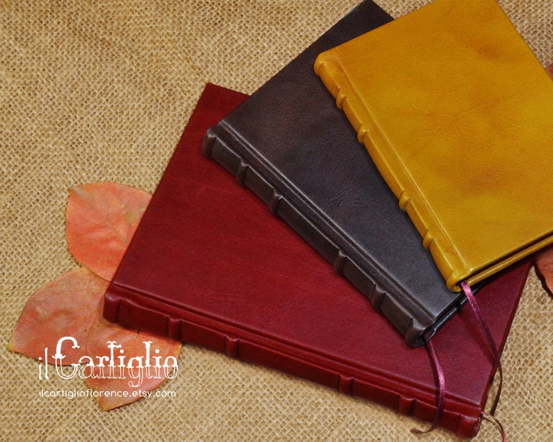 May include: Three leather-bound journals in burgundy, dark brown, and mustard yellow. The journals are stacked on top of each other and have a rustic, handmade look. The text on the bottom of the image reads "il Cartiglio ilcartiglioflorence.etsy.com".