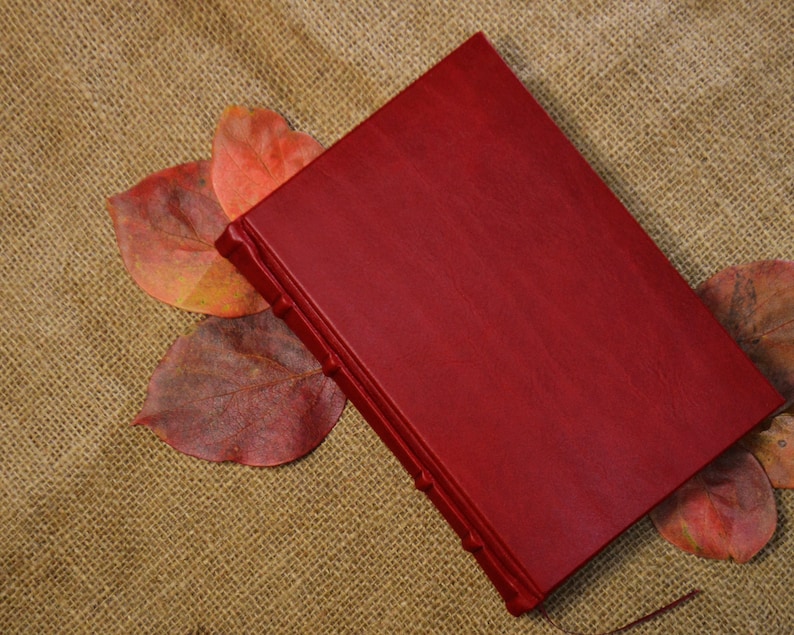 May include: A deep red leather-bound journal with a textured cover. The journal is lying on a burlap surface with several red and orange leaves scattered around it.
