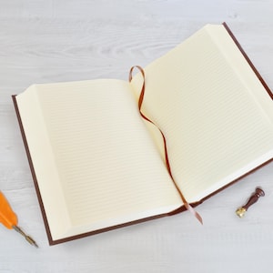 May include: An open hardback journal with cream-coloured lined pages and a dark brown cover. A thin brown ribbon bookmark extends from the centre. Beside the journal lies an orange feather quill pen with a metal nib and a small wax seal stamp.
