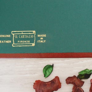 May include: A dark green leather desk pad with a reddish-brown border. The surface features gold embossed text that reads: GENUINE LEATHER, IL CARTIGLIO FIRENZE, MADE IN ITALY, enclosed within a rectangular scroll-like graphic design.
