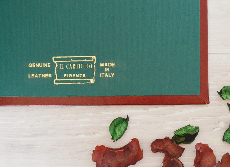 May include: A dark green leather desk pad with a reddish-brown border. The surface features gold embossed text that reads: GENUINE LEATHER, IL CARTIGLIO FIRENZE, MADE IN ITALY, enclosed within a rectangular scroll-like graphic design.