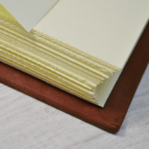 May include: A journal with a dark brown leather cover and thick, cream-coloured pages. The paper edges are rough and deckled, showing a textured, handmade quality. The book is partially open, revealing the layered stack of individual paper sheets.