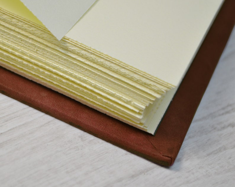 May include: A journal with a dark brown leather cover and thick, cream-coloured pages. The paper edges are rough and deckled, showing a textured, handmade quality. The book is partially open, revealing the layered stack of individual paper sheets.