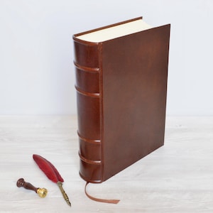 May include: A dark brown leather-bound book with a raised spine and a ribbon bookmark, accompanied by a red feather quill pen and a small metal wax seal stamp with a wooden handle, arranged together on a flat surface.