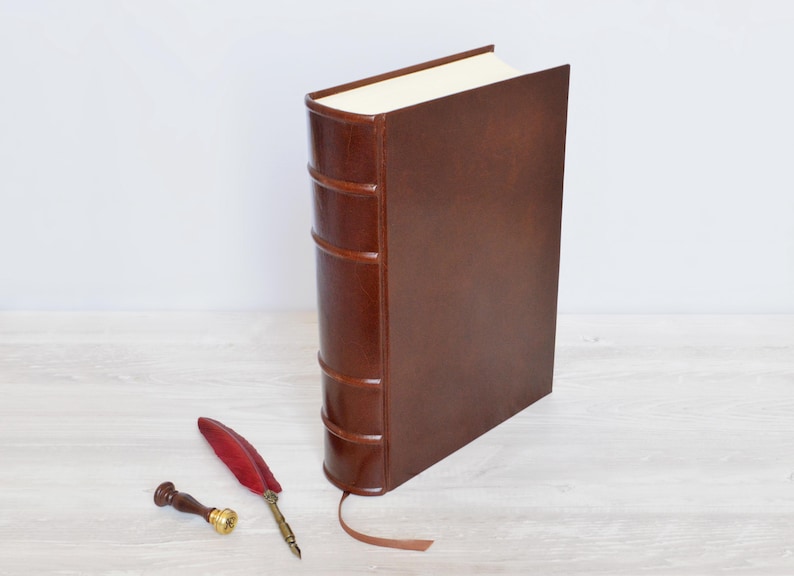 May include: A dark brown leather-bound book with a raised spine and a ribbon bookmark, accompanied by a red feather quill pen and a small metal wax seal stamp with a wooden handle, arranged together on a flat surface.