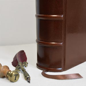 May include: A dark brown leather-bound book with a raised spine, a brown ribbon bookmark, a wooden wax seal stamp with the letter T engraved on the brass base, and a quill pen with a dark red feather and an ornate metallic handle.