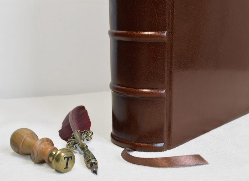 May include: A dark brown leather-bound book with a raised spine, a brown ribbon bookmark, a wooden wax seal stamp with the letter T engraved on the brass base, and a quill pen with a dark red feather and an ornate metallic handle.