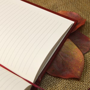 May include: A red leather-bound journal with lined pages, closed and lying on a burlap surface with two red autumn leaves.