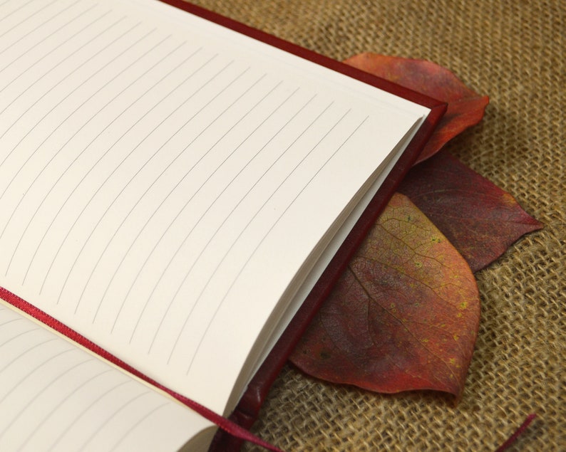 May include: A red leather-bound journal with lined pages, closed and lying on a burlap surface with two red autumn leaves.