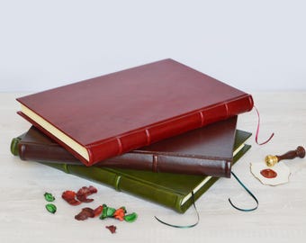 May include: A stack of three hardcover journals with smooth leather-like covers in deep red, dark brown, and olive green. Each journal features a raised spine, cream-colored pages, and a thin ribbon bookmark. A wax seal stamp and dried petals are nearby.