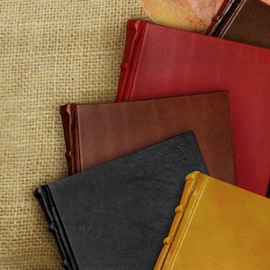 May include: Five leather-bound journals in different colors, including red, brown, black, and gold, stacked on top of each other. The journals are on a burlap background.