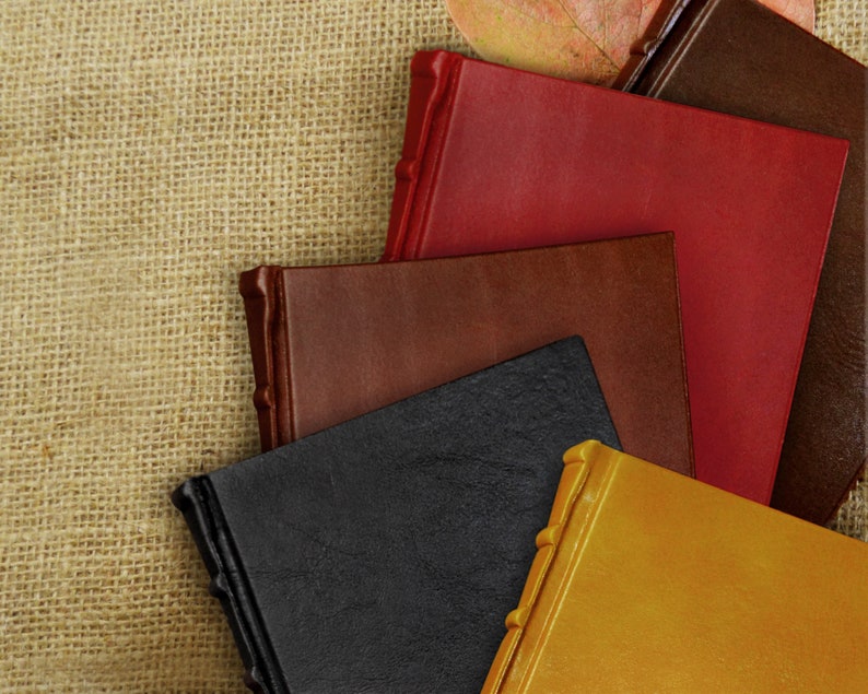 May include: Five leather-bound journals in different colors, including red, brown, black, and gold, stacked on top of each other. The journals are on a burlap background.