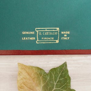 May include: A dark green leather surface featuring a gold-stamped emblem with a scroll design. The text reads: GENUINE LEATHER, IL CARTIGLIO, FIRENZE, MADE IN ITALY. A red border separates the green section from a light-colored surface below.