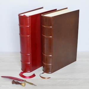 May include: Two hardback books with smooth covers in deep red and dark brown, featuring raised bands on the spines and integrated ribbon bookmarks. A red quill pen and a small wooden handle wax seal stamp are positioned in front of the books.