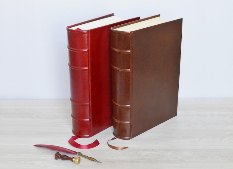 May include: Two hardback books with smooth covers in deep red and dark brown, featuring raised bands on the spines and integrated ribbon bookmarks. A red quill pen and a small wooden handle wax seal stamp are positioned in front of the books.