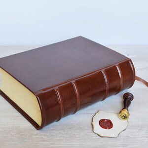 May include: A dark brown leather-bound book with a smooth finish and raised spine bands, featuring cream-colored pages and a thin brown ribbon bookmark. Beside the book lies a small brass wax seal stamp and a piece of paper with a red wax seal.