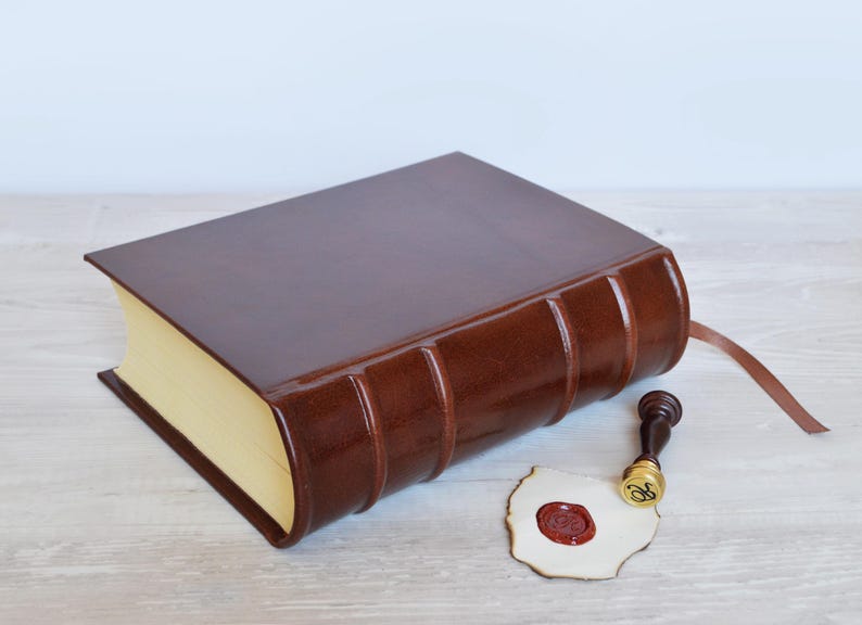 May include: A dark brown leather-bound book with a smooth finish and raised spine bands, featuring cream-colored pages and a thin brown ribbon bookmark. Beside the book lies a small brass wax seal stamp and a piece of paper with a red wax seal.