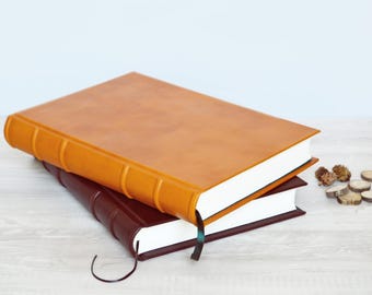 May include: Two rectangular hardcover books stacked on top of each other. The top book has a smooth tan cover with a raised spine, while the bottom book has a dark brown cover. Both books feature white pages and thin ribbon bookmarks.