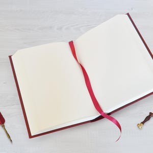May include: An open book with a deep red cover and blank cream-colored pages. A thin red ribbon bookmark extends from the center. Beside the book lies a red feather quill pen with a gold-toned metal nib and a small wax seal stamp with a wooden handle.