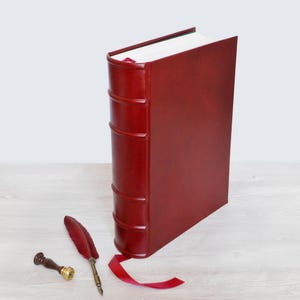 May include: A deep red leather-bound book with a raised spine and a matching red ribbon bookmark. Beside the book lies a red feather quill pen with a metallic tip and a small wooden wax seal stamp with a brass base, completing the set.