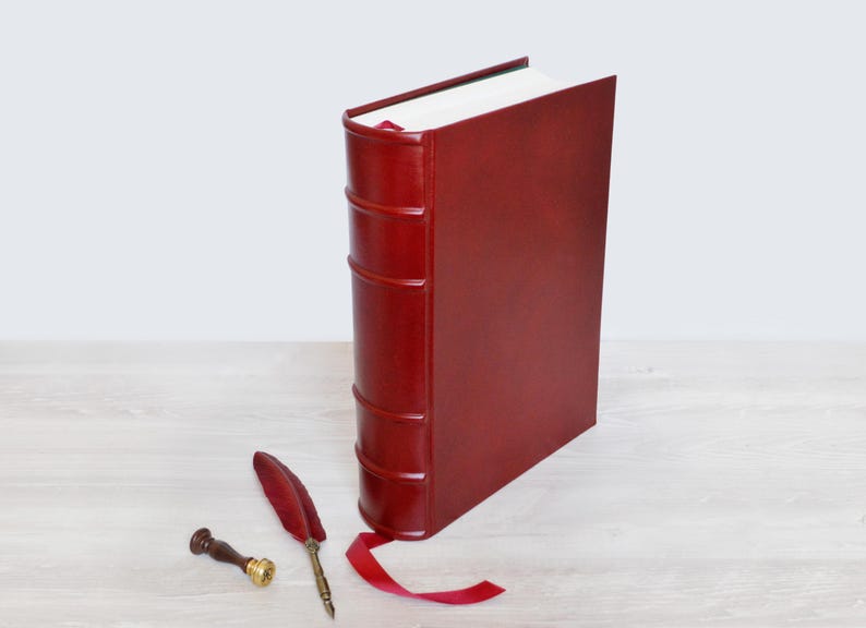 May include: A deep red leather-bound book with a raised spine and a matching red ribbon bookmark. Beside the book lies a red feather quill pen with a metallic tip and a small wooden wax seal stamp with a brass base, completing the set.
