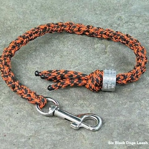 May include: A black and orange paracord dog leash with a silver tag that says "Six Black Dogs Leash Co."