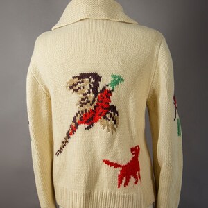 May include: A cream-colored knitted cardigan with a pheasant and a dog embroidered on the back. The pheasant is red, brown, and green, and the dog is red.