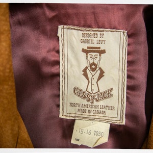 May include: A brown leather jacket with a burgundy lining and a label that reads "Designed by Gabriel Levy Gassy Jack North American Leather Made in Canada 15-16 7050 Size".