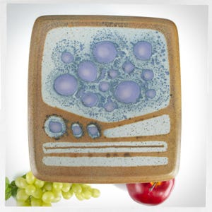 May include: A square ceramic plate with a light blue glaze and a pattern of purple and blue circles. The plate has a brown rim and a brown design around the edge.