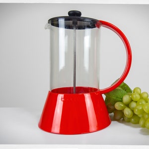 Peut inclure: Une cafetière à piston en verre et plastique rouge avec une carafe en verre transparent et un couvercle noir. La carafe a une base rouge et la poignée est rouge. Un bouquet de raisins verts est en arrière-plan.