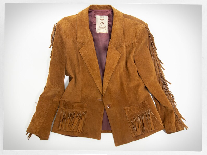 May include: A brown suede jacket with fringe detailing on the sleeves, pockets, and hem. The jacket has a single button closure and a burgundy lining.