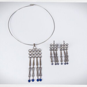 Puede incluir: Un conjunto de collar y pendientes plateados. El collar tiene un collar circular r&iacute;gido con un colgante con un dise&ntilde;o geom&eacute;trico con detalles azules. Los pendientes reflejan el dise&ntilde;o del colgante, con elementos colgantes y cuentas azules.