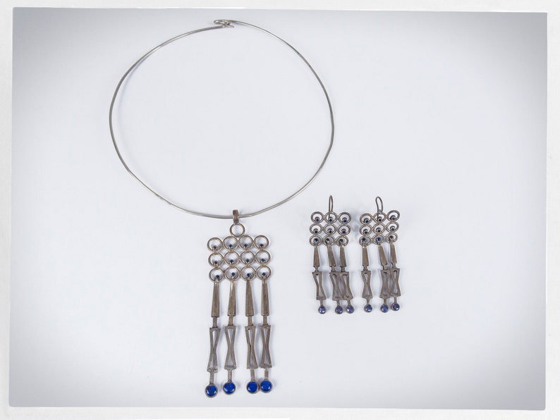 Puede incluir: Un conjunto de collar y pendientes plateados. El collar tiene un collar circular r&iacute;gido con un colgante con un dise&ntilde;o geom&eacute;trico con detalles azules. Los pendientes reflejan el dise&ntilde;o del colgante, con elementos colgantes y cuentas azules.