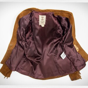 May include: A brown suede jacket with a burgundy satin lining. The jacket has fringe detailing on the sleeves and bottom hem. The label inside the jacket reads "Suede Leather Jacket".