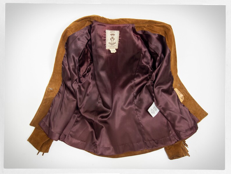 May include: A brown suede jacket with a burgundy satin lining. The jacket has fringe detailing on the sleeves and bottom hem. The label inside the jacket reads "Suede Leather Jacket".
