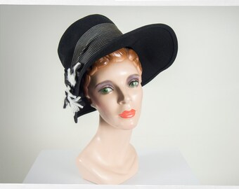 May include: A black felt hat with a wide brim and a white and black trim. The hat is angled slightly to the side and has a decorative bow on the side.