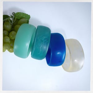 May include: Four plastic bangles in shades of blue and green. The bangles are stacked on top of each other. The top bangle is a light green, the second is a teal blue with white swirls, the third is a dark blue, and the bottom bangle is a translucent white.