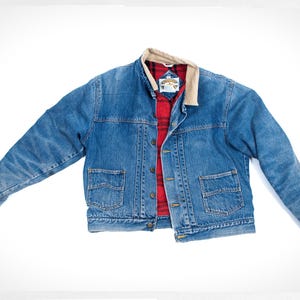 May include: A blue denim jacket with a red and black plaid lining and a tan collar. The jacket has two front pockets and a button-up closure. The jacket is laid flat on a white background.