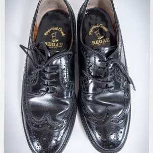 Op de afbeelding: Een paar zwarte leren brogueschoenen met een wingtip-ontwerp. De schoenen hebben een gouden label aan de binnenkant met de tekst "Imperial Grade Regal Since 1904".