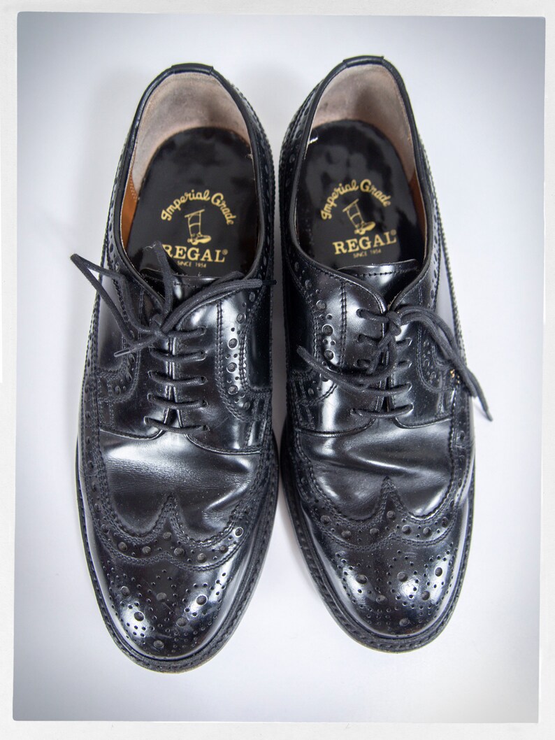 Op de afbeelding: Een paar zwarte leren brogueschoenen met een wingtip-ontwerp. De schoenen hebben een gouden label aan de binnenkant met de tekst "Imperial Grade Regal Since 1904".
