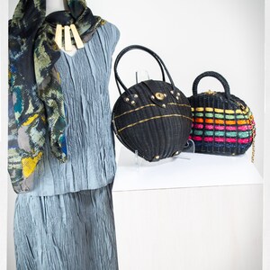 May include: A blue pleated dress with a black and white patterned scarf. Two round woven handbags, one black and one with rainbow stripes, are displayed on a white surface.