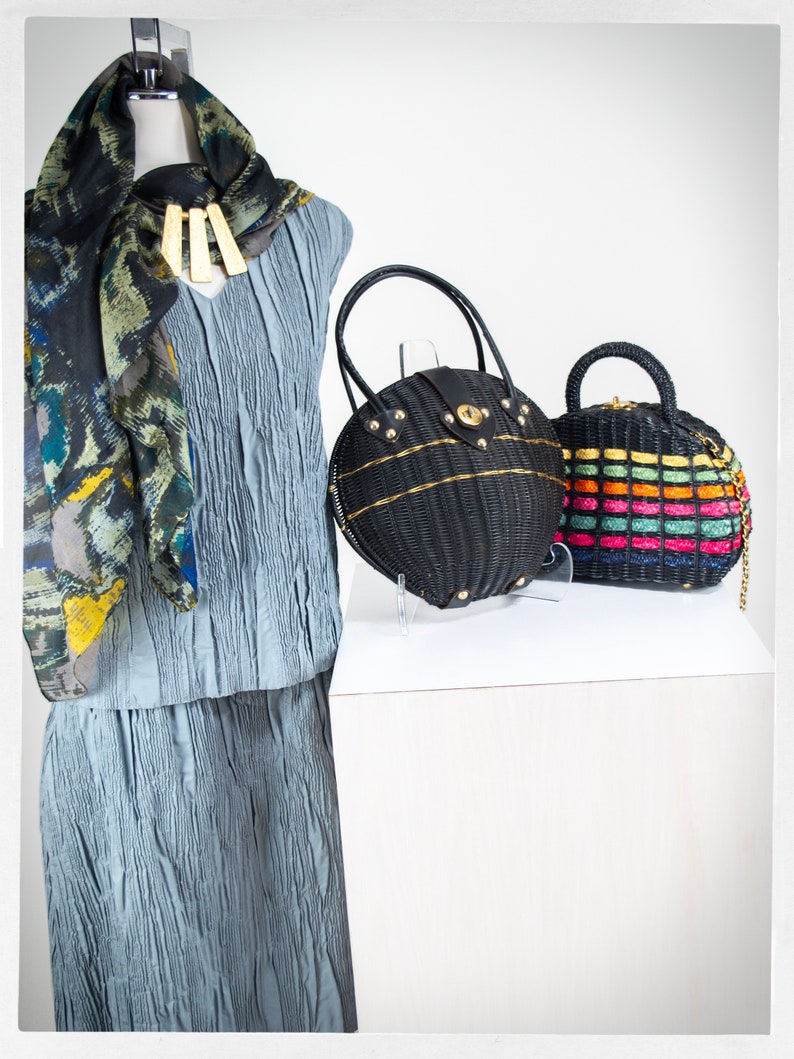 May include: A blue pleated dress with a black and white patterned scarf. Two round woven handbags, one black and one with rainbow stripes, are displayed on a white surface.