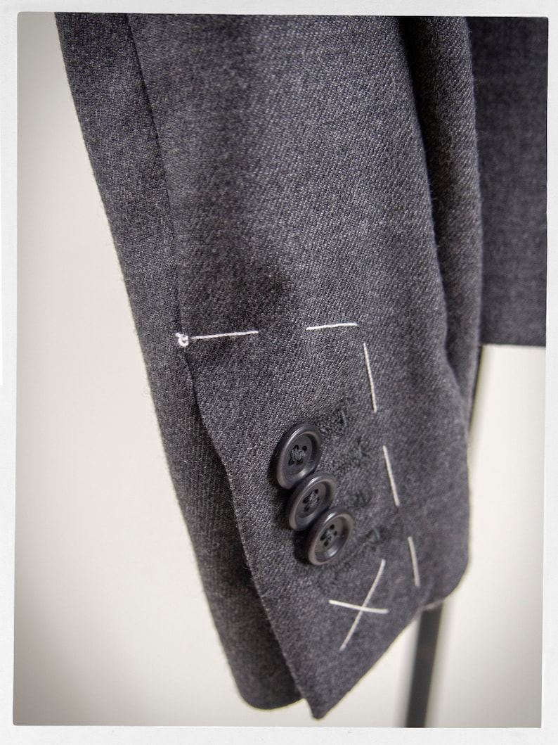 May include: A close-up of a dark gray blazer sleeve with three black buttons and white stitching.
