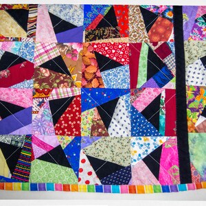 May include: A colourful patchwork quilt featuring a range of fabrics and patterns. The quilt is made up of squares and triangles, and it is bordered with a rainbow-coloured trim.